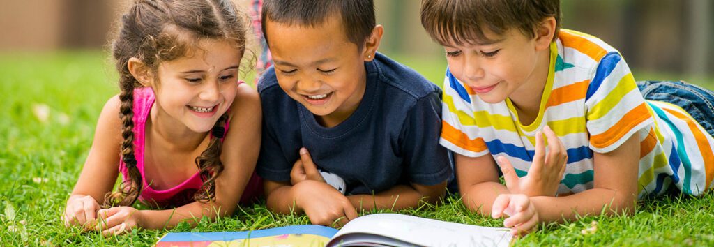 three Children reading books on green grass.